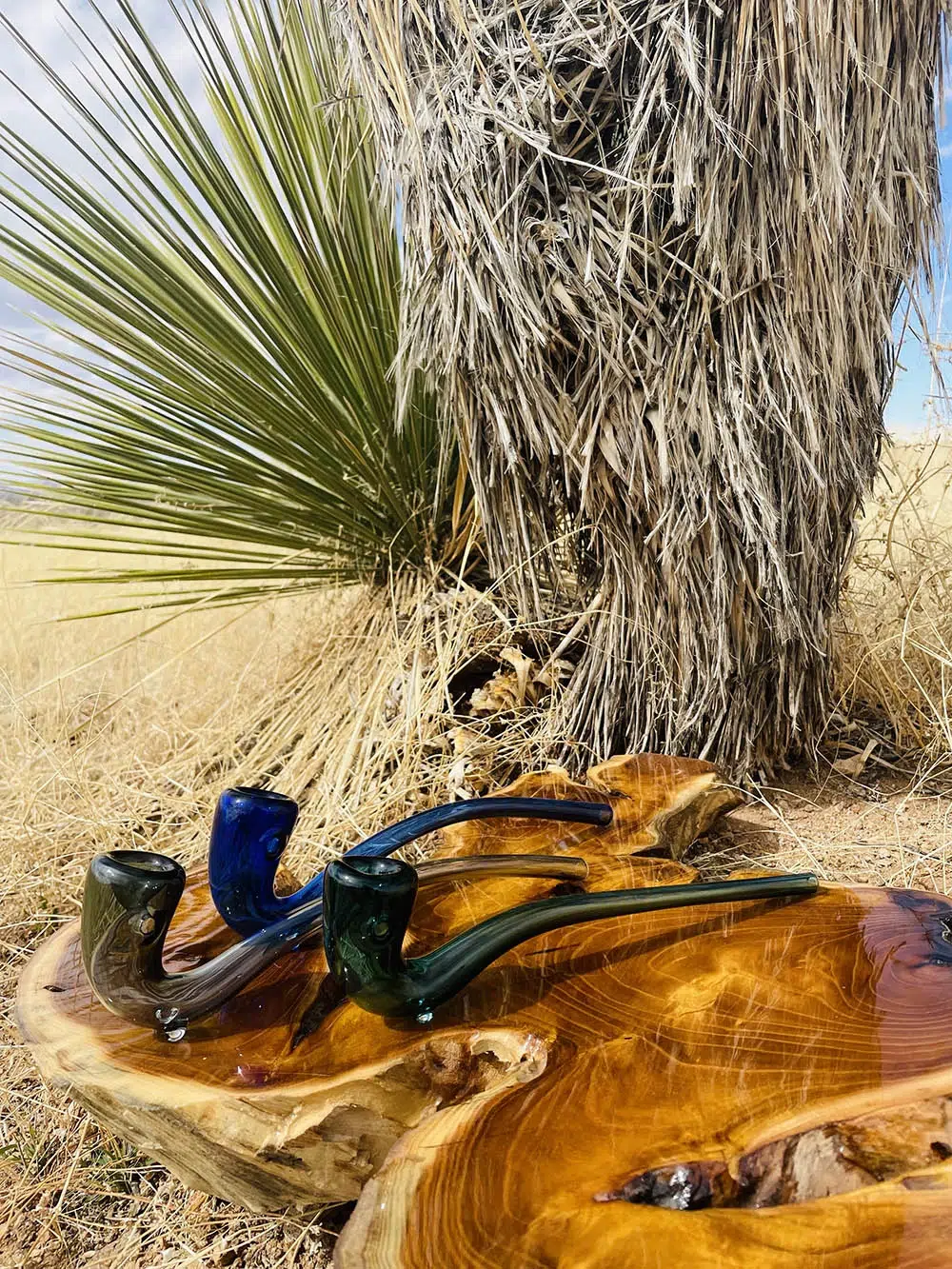 three staggered glass gandalf pipes in teal black and blueon wooden table with cactus palm and yellow brush behind