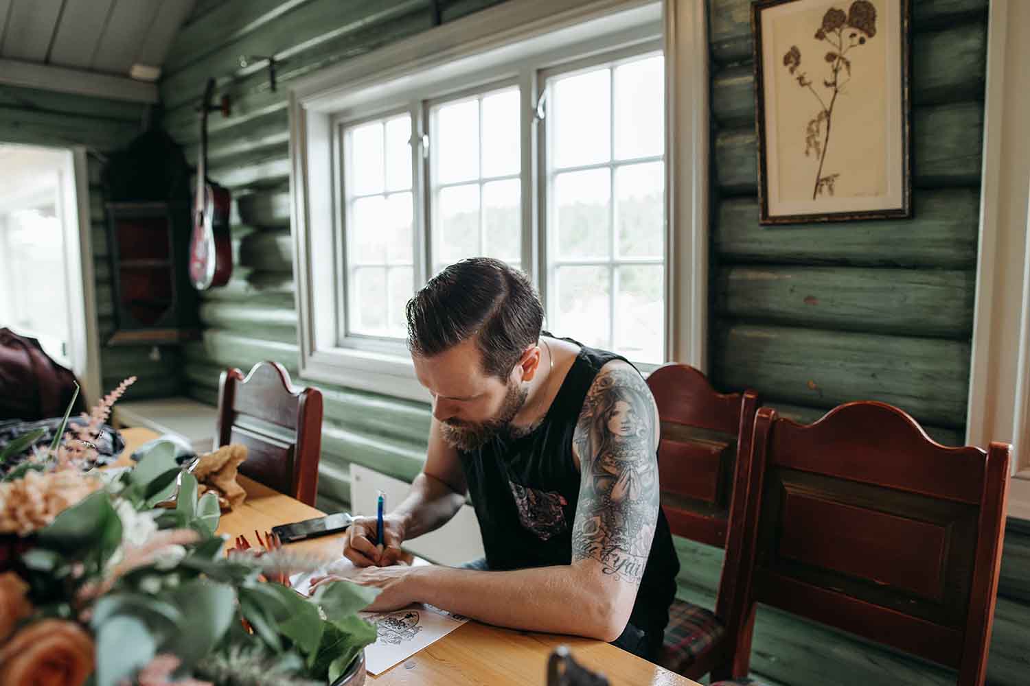 man with tattoos drawing cartoons on wooden table in living room with plants around and window behind