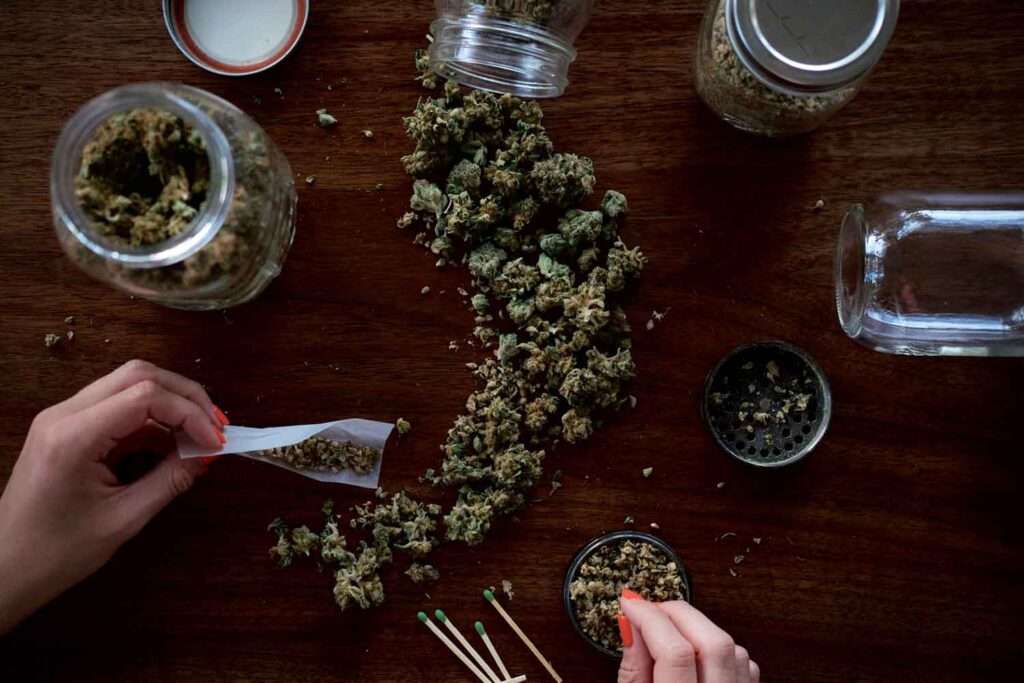 an ounce of weed spread across table with mason jars and a woman rolling a joint